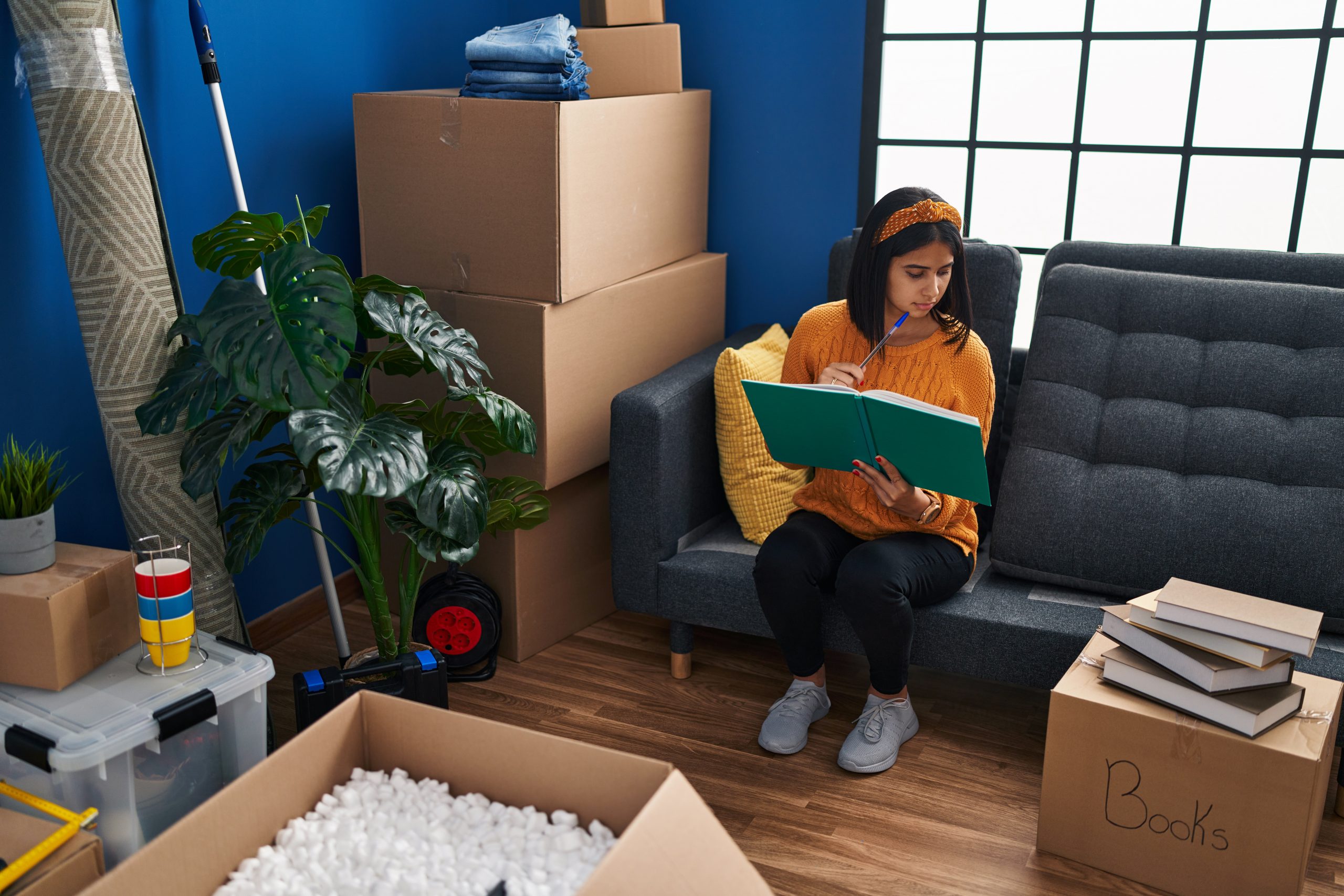Young latin woman reading book sitting on sofa at new home packing boxes for removals